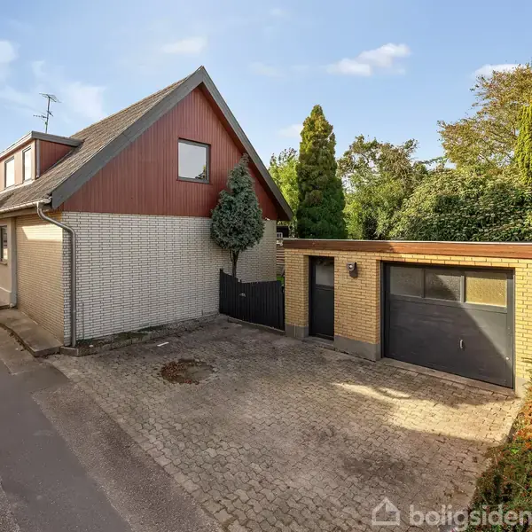 Et hvidt og rødt teglhus med en sort garageport står ved en vejkant. Ved siden af er der en garage. Omkring huset er der buske og træer.
