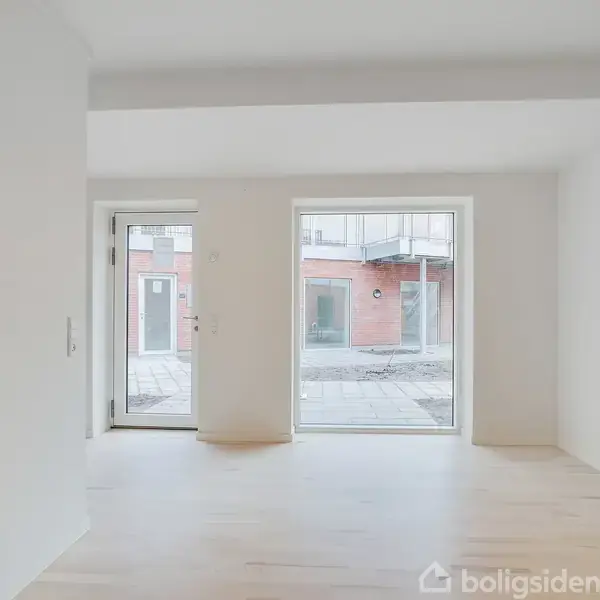 Et tomt værelse med lyse trægulve har to store vinduer og en glasdør. Udenfor ses en gårdhave og en rød murstensbygning med vinduer og balkoner.