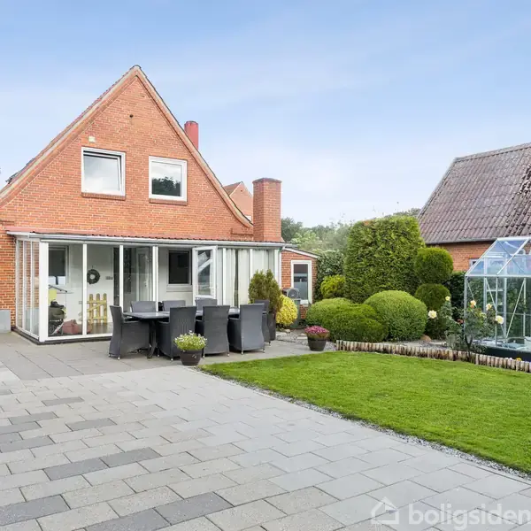 Rødstenshus med stort skråt tag, har en udestue og terrasse med sorte møbler. På plænen er der et drivhus omgivet af velplejede buske og flisebelægning.