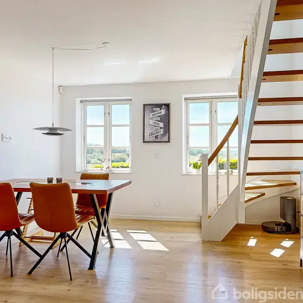 Spisebord med fire orange stole står i en lys stue med trægulve. En moderne trappe med trætrin fører opad. Store vinduer giver udsigt og lysindfald.