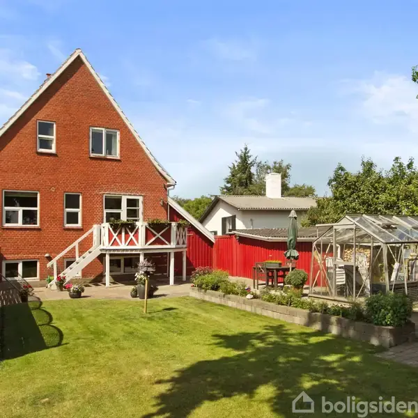 Rød murstensvilla med hvidrammede vinduer står i en grøn, velholdt have. Et drivhus og et bord med parasol ses ved siden af huset, omkranset af træhegn.