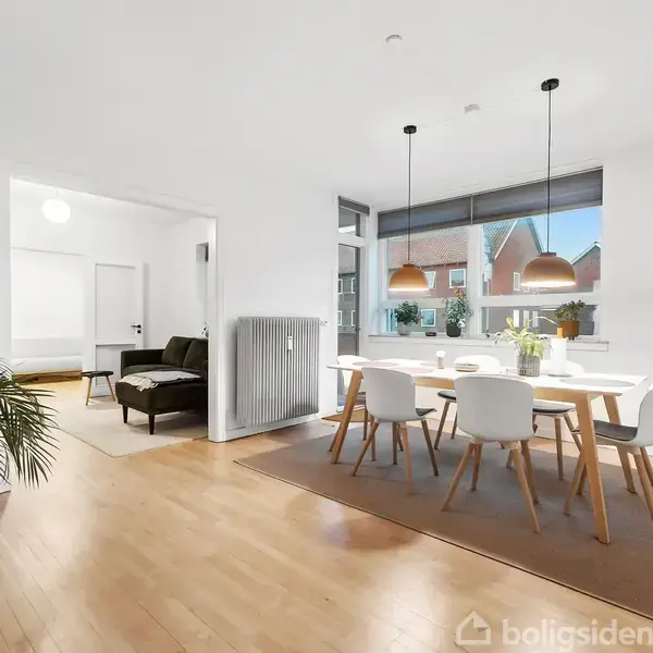 Et moderne spisestueopstilling med et lyst træbord og seks hvide stole. Rummet har store vinduer med udsigt, hængende lamper ovenover, og en åben forbindelse til en stue med en sofa. En plante står til venstre, og en hylde med dekorationer er placeret i hjørnet.