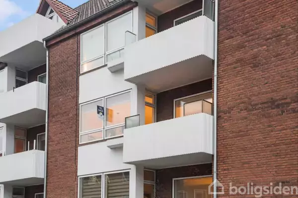 Murstensbygning med hvide altaner og store vinduer, set fra gadeniveau. På en altan hænger et "til salg" skilt. Bygningen er delvist oplyst indefra.