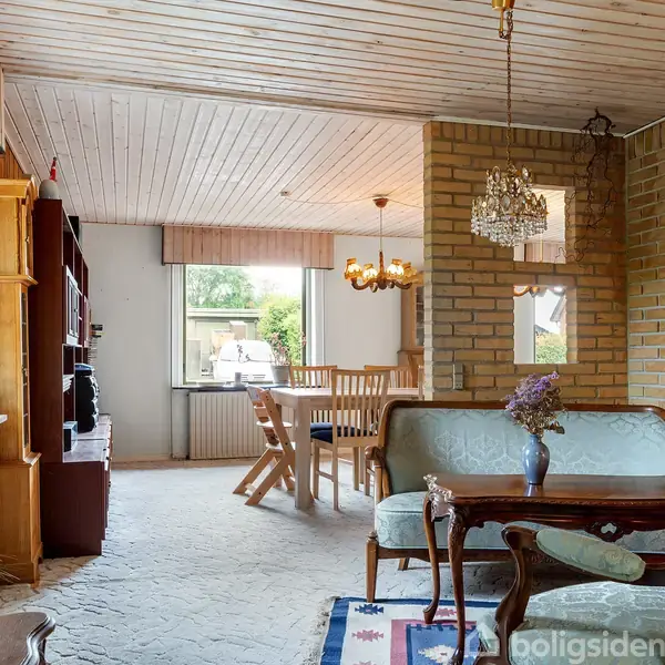 Stue med klassisk møblement, sofa og sofabord i forgrunden. Spisebord med stole i baggrunden. Lysindfald fra vindue, træloft og murstensvægge. Lamper hænger fra loftet.
