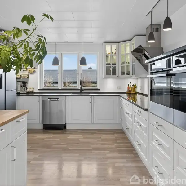 Køkken med hvide skabe og indbygget ovn. Plante på hylde til venstre. Vinduet for enden giver udsigt over landskabet. Moderne apparater og lyse trægulve dominerer rummet.