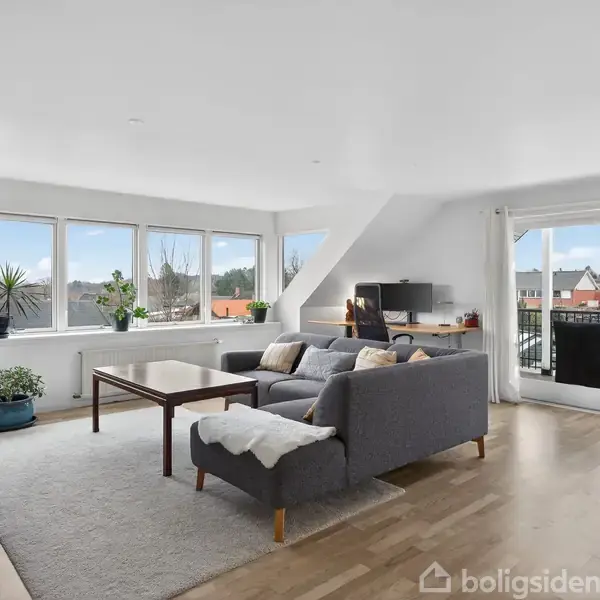 Gråt sofaarrangement står stille i en lys stue med store vinduer. Rummet har trægulv, grønne planter og adgang til en altan med udsigt over et nabolag. Ingen tekst fundet.