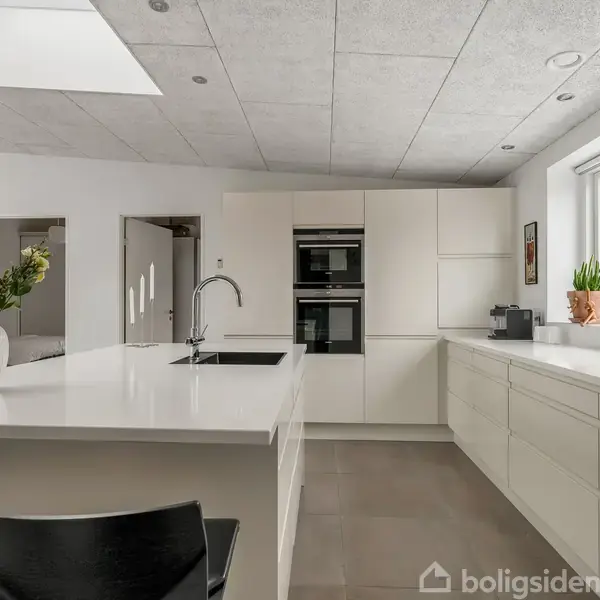 Et moderne køkken med en stor hvid køkkenø, hvorpå står en vase med blomster. Der er integrerede ovne i en hvid skabsvæg. Store vinduer giver lysindfald.