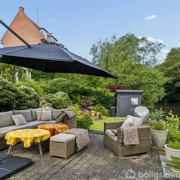 En have med sofaarrangement under en stor parasol. Sofaen og borde er placeret på en flisebelagt terrasse omgivet af grønne buske og træer nær en sø.