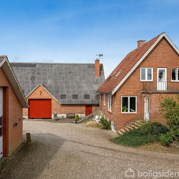 Et rødt murstenshus står med en stejl gavltag ved siden af en garage og ladebygning. Gårdspladsen er belagt med grus, og der er grøn beplantning i forgrunden.