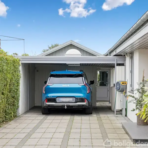 Blå bil parkeret i en carport ved siden af et hvidt hus. Flisebelagt indkørsel omgivet af grønne hække og planter under en blå, delvist skyet himmel.