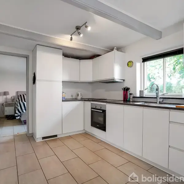 Et moderne køkken med hvide skabe og skuffer, integreret ovn og kogeplade. Et vindue ved siden af vasken giver udsigt til haven. Til venstre ses en døråbning.