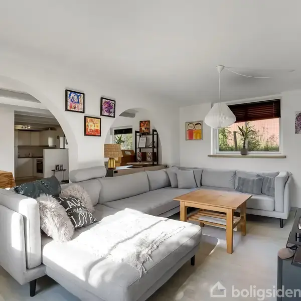 En stor grå hjørnesofa står i en lys stue ved siden af et træsofabord. På væggene hænger farverige malerier. I baggrunden ses et spisebord og et åbent køkken.