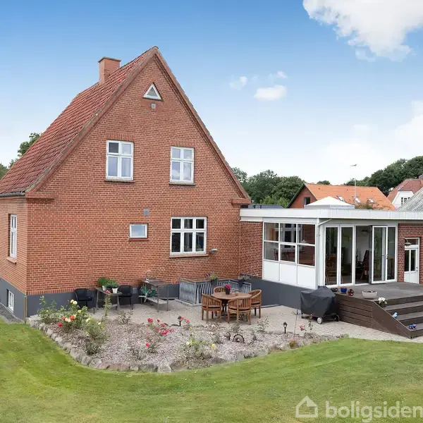 Rødt murstenshus står i solen, omgivet af en velplejet have med blomsterbede. På terrassen står et rundt havebord med stole. Huset har en moderne tilbygning med glaspartier.