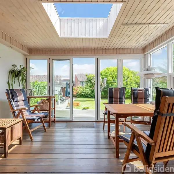 Et lyst udestueinteriør med træmøbler, herunder stole og et bord. Store vinduer og et ovenlys slipper masser af naturligt lys ind. Udsigt til en grøn have udenfor.