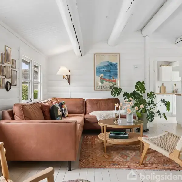 En stue indeholder en brun lædersofa, en tæppebelagt gulv, planter på et rundt sofabord, hvide trævægge og loftsbjælker. Væggene har kunst og lyse belysningsarmaturer.