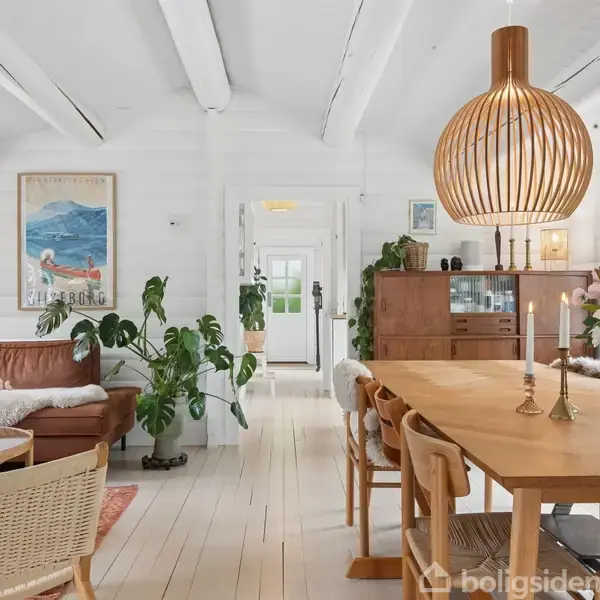 Stue med sofa og spisebord med tændte stearinlys. Væggene er hvide, og rummet er lyst med store vinduer. En stor loftlampe hænger over bordet, og planter pynter.