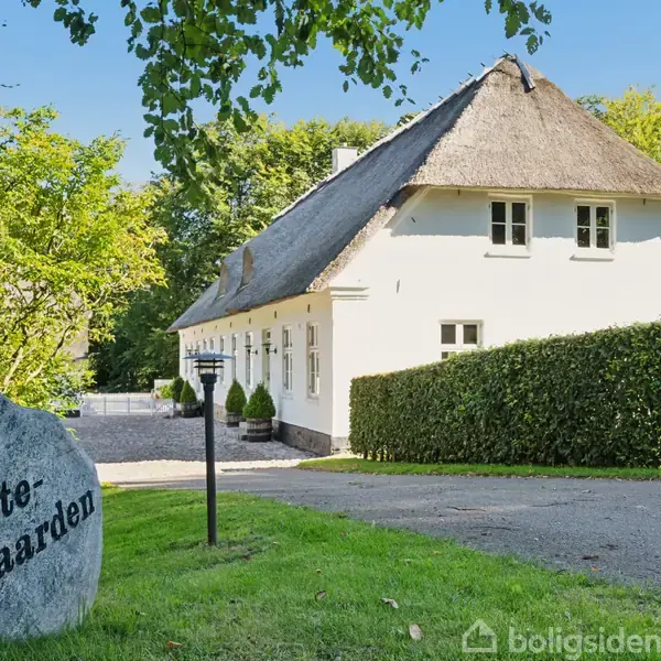 En hvid, stråtækt hus med vinduer står i en frodig have med buske og træer. En stenmarkeret indkørsel bærer teksten "Præstegaarden".