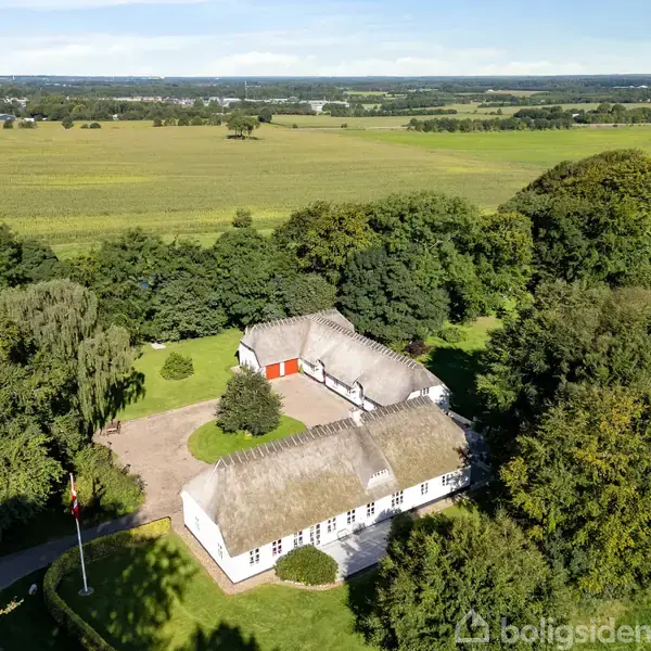 Bindingsværkshus med stråtag står stille omgivet af tæt skov, marker og en stor græsplæne. En cirkulær indkørsel omgiver huset, mens flagstangen står ved siden af en indgang.