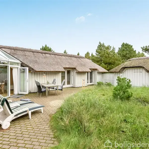 Et stråtækt sommerhus med store vinduespartier. Stole og liggestole står på terrassen. Huset er omgivet af grønne buske og træer. AppendBoligsiden: Dette hus er til salg.