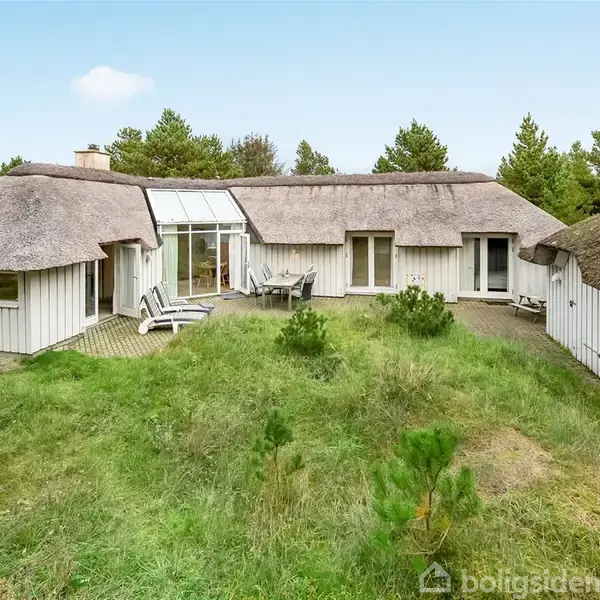Stråtægt hus med store vinduer, står omgivet af grønne buske og træer. Hvidmalede terrassemøbler på flisebelagt terrasse. Klar himmel ses i baggrunden.
