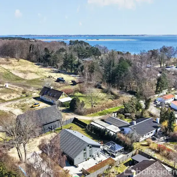 En gul bil holder stille ved et hus omgivet af grønne træer og vådområder. I baggrunden ses en skov og en klar blå sø, der skaber en fredelig landskabsudsigt.