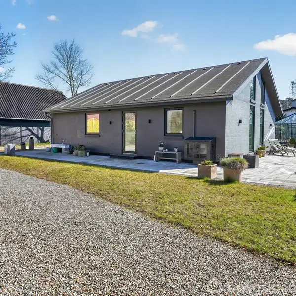 Et moderne hus med ensidet tag står omgivet af en grusvej og have. En træterrasse med møbler ses til højre, mens et drivhus kan skimtes i baggrunden.