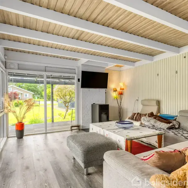 Stue med stor hjørnesofa, der vender mod et sofabord og fladskærms-tv på væggen. Glasdøre åbner til have med græsplæne og træer. Skjulte lamper skaber hyggelig belysning.