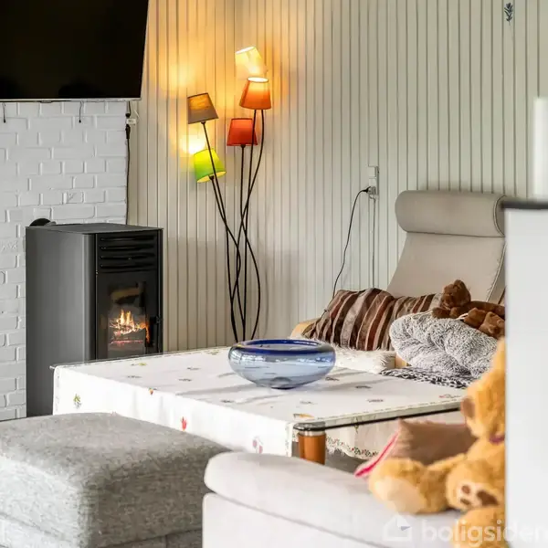 Brændeovn opvarmer en stue med en lænestol, sofabord og pyntelamper. Et tæppe, puder og en skål står på bordet. Området er hyggeligt indrettet med varme farver.