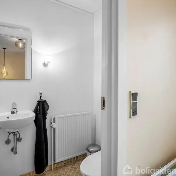 En lille badeværelse med håndvask og spejl på væggen, to lys over spejlet, et sort håndklæde ved siden af en radiator og en toiletstol i et lyst rum.