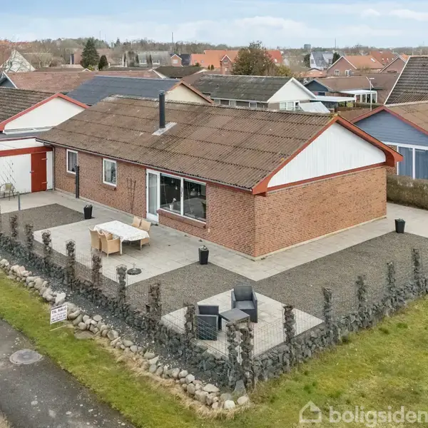 En rød murstensbungalow med en stor, flisebelagt terrasse omgivet af en lav murstensmur; havemøbler er placeret på terrassen. Omgivet af nabohuse i et villakvarter.