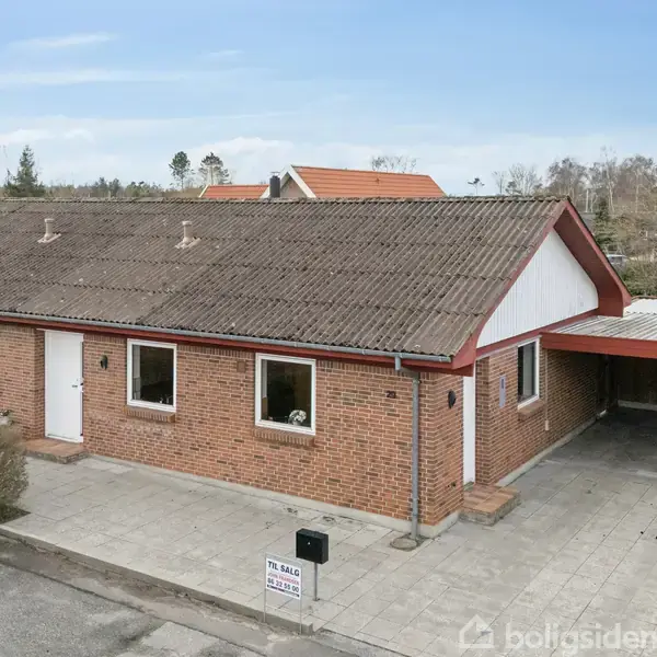 Et rødt murstenshus med skråt tag og carport. Huset er omgivet af en flisebelagt forhave. Et skilt ved indkørslen viser teksten "Til salg" og kontaktoplysninger.