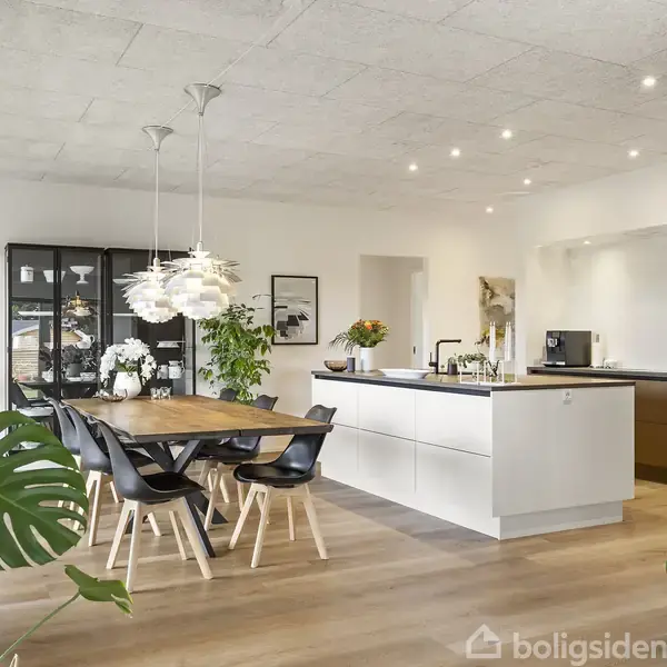 Et moderne køkken-alrum med hvidt køkkenbord, træspisebord og sorte stole. Planter og belysning skaber en lys og stilfuld atmosfære i det åbne rum.