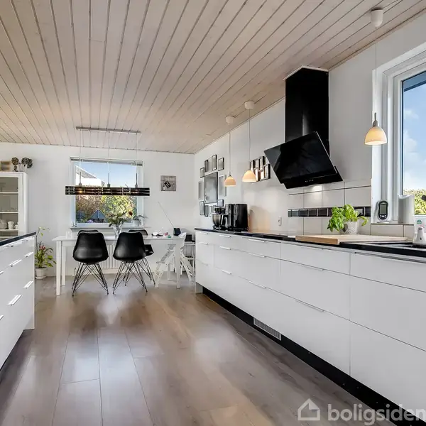 Køkken med hvide skabe og sort emhætte, dagslys strømmer ind fra stort vindue, spisestue med sorte stole og bord i baggrunden. Moderne indretning med træloft og planter.