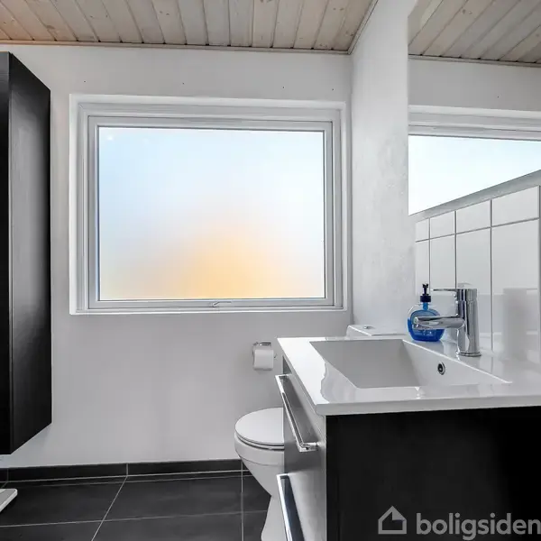 Et moderne badeværelse med flisegulv. En vask med sæbedispenser står på et skab. Et toilet er placeret ved siden af, og der er et stort vindue med mat glas.