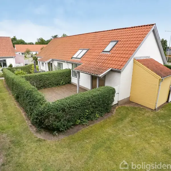 Et rødt tegltagshus med en lille veranda omkranset af en hæk. Huset ligger i en have med græsplæne og er omgivet af andre lignende boliger.