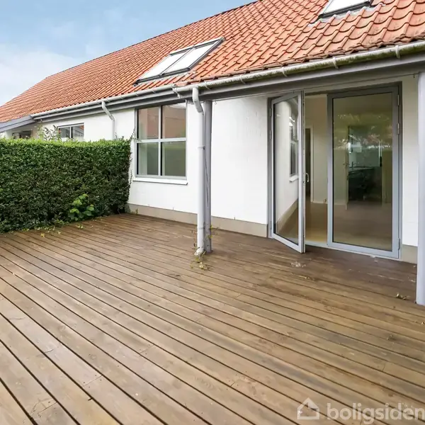 Terrasse med åbne glasdøre, der fører til et indre rum. Omgivet af en hæk og udsigt til et rødt tegltag. Ingen tekst fundet.