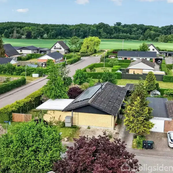 Boligkvarter med huse omgivet af træer og haver. En vej løber gennem området, mens åbne marker og skov ses i baggrunden på en solrig dag.