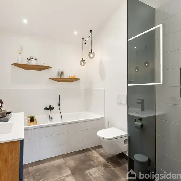 Et moderne badeværelse med et fritstående badekar og vægmonteret toilet. En håndvask med træskab står til venstre. Væghylder og plantede, runde lamper skaber en hyggelig atmosfære.
