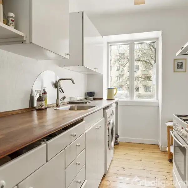 Et lyst køkken med hvidt interiør og træbordplade. En vask er indbygget i bordpladen, og der er en ovn til højre. Et vindue giver naturligt lysindfald.