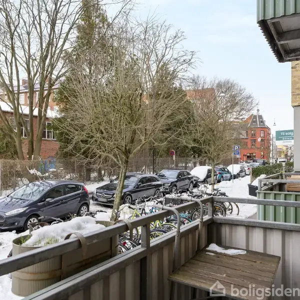 En lille altan med træbord har sne på rækværket. I baggrunden ses en sneklædt gade med parkerede biler, cykler og træer langs fortovet.
