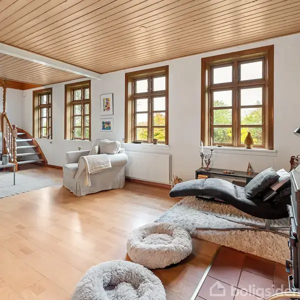 Et lyst stueområde med trægulv og loft, møbleret med sofaer og en chaiselong. Trætrappe i hjørnet og store vinduer med udsigt til grønne omgivelser.