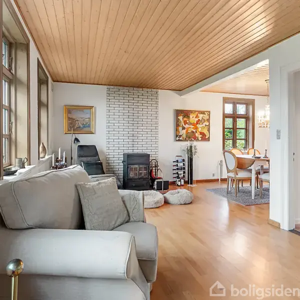 Gråt sofaarrangement står i en lys stue med parketgulv og hvidmalede vægge. En brændeovn er placeret mod en hvid murstensvæg ved siden af spiseområdet, indrammet af stort vindue.