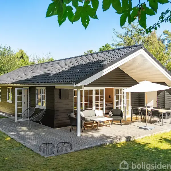Sort træhus med terrasse har havemøbler og parasol. Sollys skinner på græsplæne omkring huset, omgivet af træer og buske.