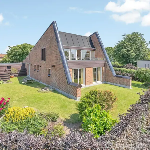 En moderne murstensvilla står i en velplejet have. Huset har unikke skråtstillet tagkonstruktioner og store vinduer. Farverige blomsterbede og en tæt hæk omkranser grunden under en klar himmel.