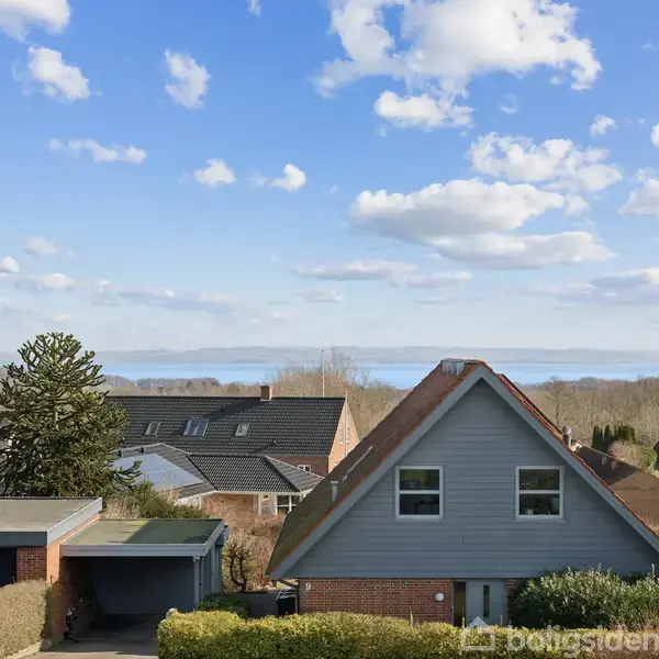 Et gråt hus med en spidsgavl står stille blandt andre huse i et boligområde med udsigt til havet i det fjerne under en blå, skydækket himmel.