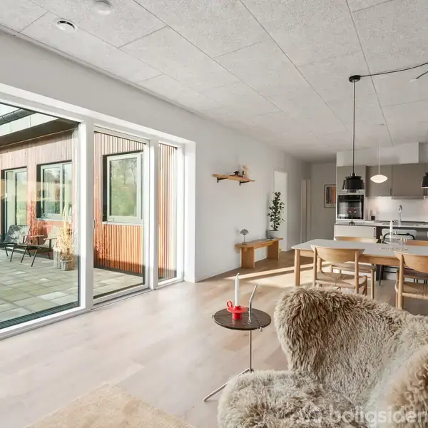 En moderne stue med spisebord og stole, set mod et åbent køkken med hængelamper. Gennem store vinduer ses en terrasse med havemøbler og udsigt til en have.