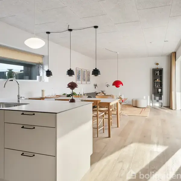 Køkken med hvidt skab og vask, omkranset af spiseområde med træmøbler. Moderne belysningsarmaturer hænger fra loftet. Store vinduer til højre giver naturligt lys.