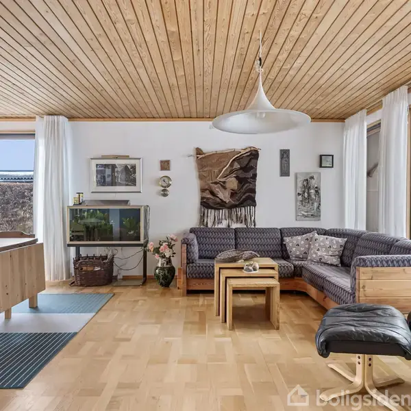 En L-formet sofa står i en lys stue med trægulve og træloft. Ved siden af er et spisebord. Store vinduer med hvide gardiner slipper lys ind. Vaskebalje med planter og vægbilleder skaber hygge.