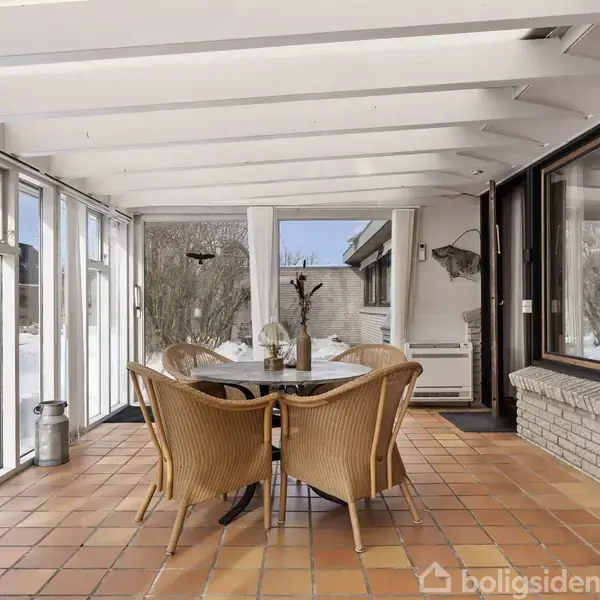 Et rundt bord omgivet af fire fletstole står på gulv af terracottafliser i en lys havestue, hvor store vinduer giver udsigt til en sneklædt have.