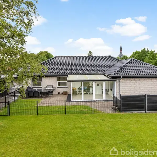 En moderne villa står stille omgivet af en grøn græsplæne og hegn; terrassen er fyldt med havemøbler, mens træer og en blå himmel skaber en rolig atmosfære.